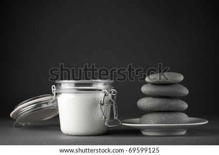 Glass jar of cosmetic cream and stack of massage stones on dark grey background.