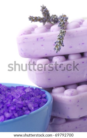 Close-up of violet bath salt in blue bowl and stack of lilac soap with twigs of lavender on white background.
