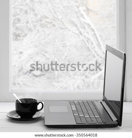 Cozy winter still life: laptop and cup of hot coffee on windowsill against snow landscape from outside.