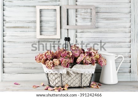 Shabby chic still life: bunch of vintage pink dry roses in wire basket and jug against white wooden blinds with empty photoframes.