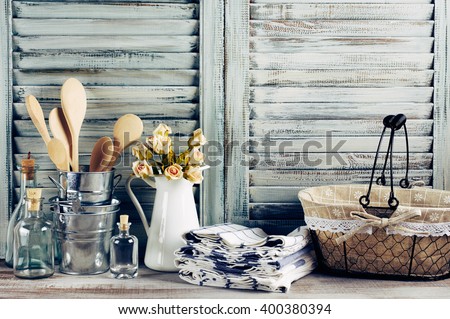 Rustic kitchen still life: wire basket, galvanized buckets with wooden spoons, jug with roses bunch, towels stack and glass bottles against vintage wooden shutters. Filtered toned image.