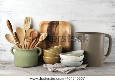 Simple rustic kitchenware against white wooden wall: rough ceramic pot with wooden cooking utensil set, stacks of ceramic bowls, jug and wooden trays.