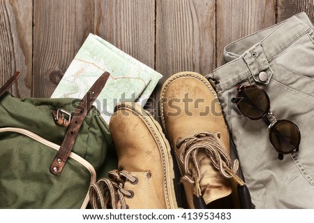 Travel accessories set on wooden background: old hiking leather boots, pants, backpack, map and sunglasses. Top view point.