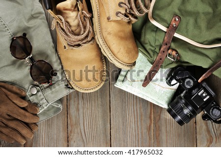 Travel accessories set on wooden background: old hiking leather boots, pants, backpack, map, gloves, vintage film camera and sunglasses. Top view point.