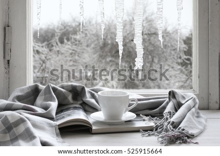Cozy winter still life: cup of hot coffee and opened book with warm plaid on vintage windowsill against frozen big icicles from outside..