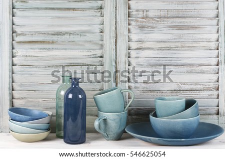 Simple rustic kitchen still life: handmade blue ceramic dish, bowls, mugs and glass bottles against shabby wooden shutters.