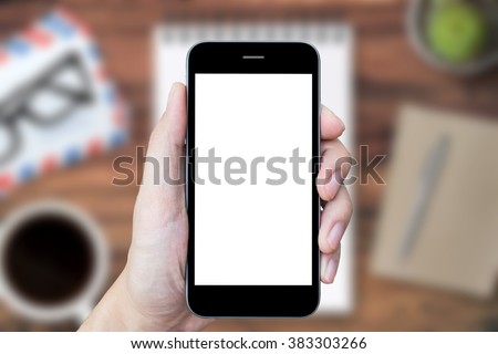 Hand holding a smartphone with white blank mockup screen on blurry wood office desk as a background.