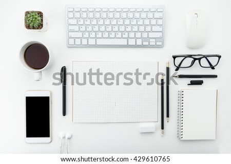 A white modern business desk table with IT gadgets and office supplies on it. Top view. The blank notebook page in the middle can be put some texts on it.
