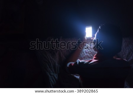 Asian young man using his mobile phone on the bed in dark room.\
Thin guy playing his smartphone on the bed in dark room.