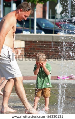 Man stopping water with foot in city park with boy toddler covering face in anticipation of water squirting up.