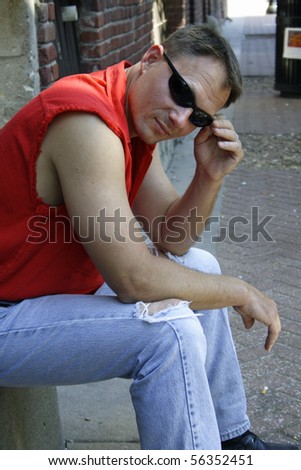 Sexy male biker sitting on curb of old street wearing sunglasses.