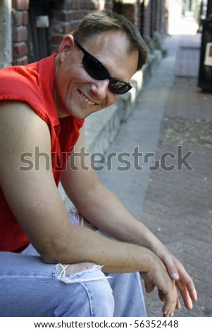Sexy male biker sitting on curb of old street wearing sunglasses and smiling.
