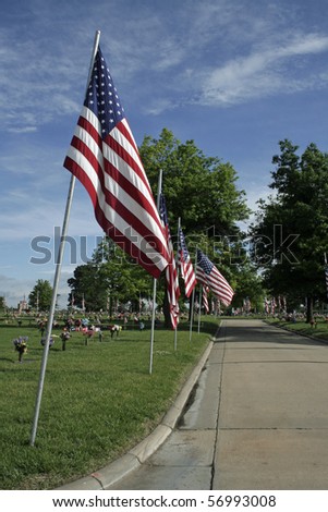American flags lining street through cemetery with cloud streaked blue sky in background.