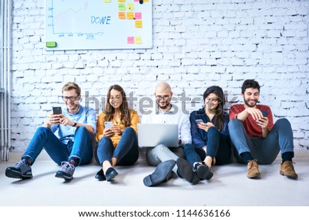 Group of university students sitting on floor together during break and using smartphones or laptop