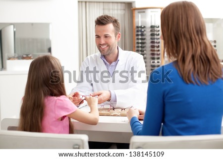 Optician, optometrist giving glasses to try for young girl visiting optical shop with her mother