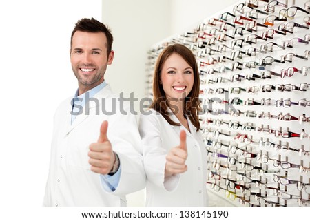 Two happy opticians, optometrists showing thumbs up in optical shop