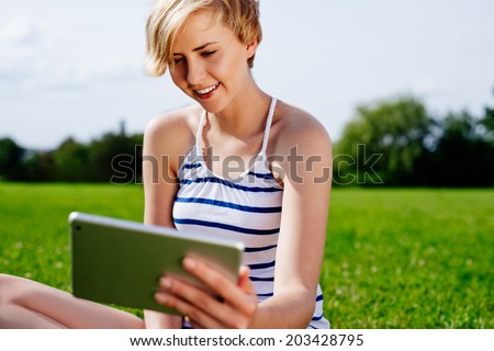 Photo of a young woman sitting on the grass and browsing the web on her tablet