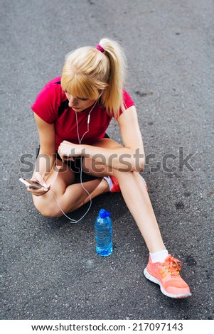 Young pretty runner sitting on the ground and choosing songs to keep her company during the run
