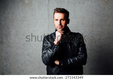 Handsome thoughtful man in leather jacket standing against concrete background looking at camera
