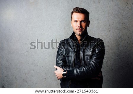Confident handsome young man in leather jacket standing against concrete wall