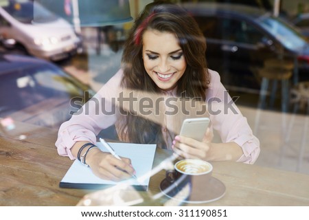 Female student at cafe working with smartphone and notepad, writing down ideas