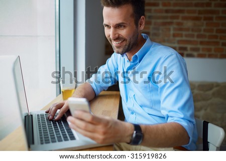 Happy mid adult man paying for online shopping with smartphone, typing on laptop computer