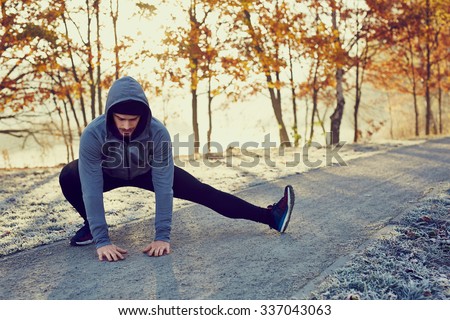 Male runner doing stretching exercise preparing for morning running workout in the park during sunny and cold fall