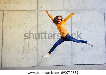 Joyful young lady jumping and raising arms in front of wall outside