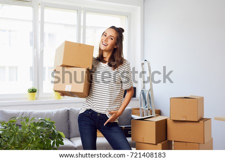 Smiling young woman moving to new apartment, holding boxes and looking at camera