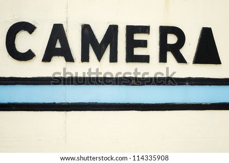 Painted camera sign with stylized black lettering above blue and black stripes