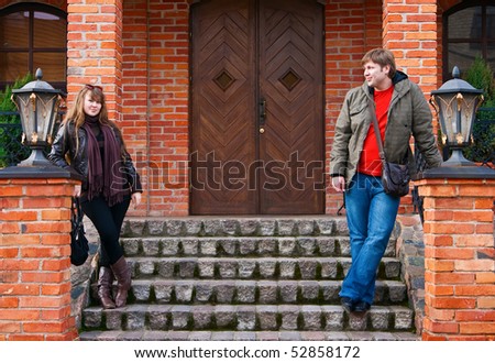 young couple standing on a steps near an old house with lamps, he is looking on her, she is looking straight and smiling