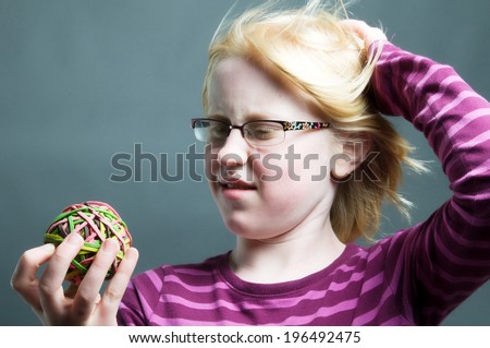 A blonde haired girl with glasses looking at a rubber band ball.