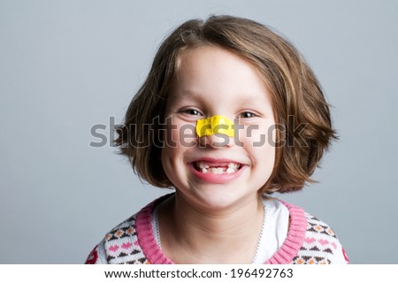 A smiling, brunette girl with missing teeth and a yellow bandage on her nose.