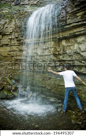 A man in a white t-shirt is holding his hand under a waterfall.