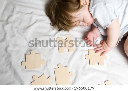 A toddler asleep on a bed surrounded by large wooden jigsaw puzzle pieces.