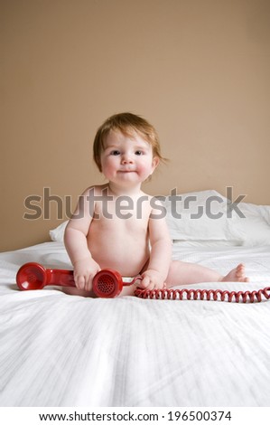 A baby sitting on a bed, holding a red corded phone horn.