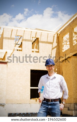A man with a builders helmet standing in front of a wooden, half built house.