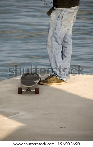 A person in jeans standing next to a skateboard at the water\'s edge.