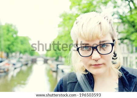 A blond haired girl with glasses and a pierced nose ring standing near a river.