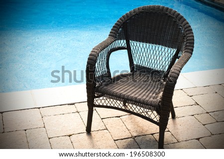 A dark wicker chair sits on a stone patio beside a pool.
