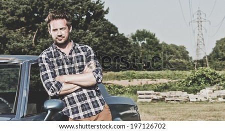 A man in a checked shirt leaning against a car.