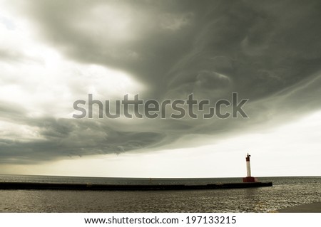 A very dark cloud above a light house and large body of water.