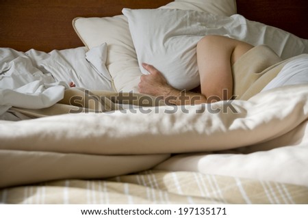 A person covering their head with a pillow while lying in bed.