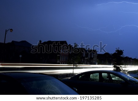 A road bordering dark houses with lightning in the background.