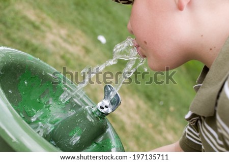 A boy drinking from a green water fountain in a park.