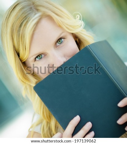 A woman with blond hair and green eyes covers her mouth with a blue book.