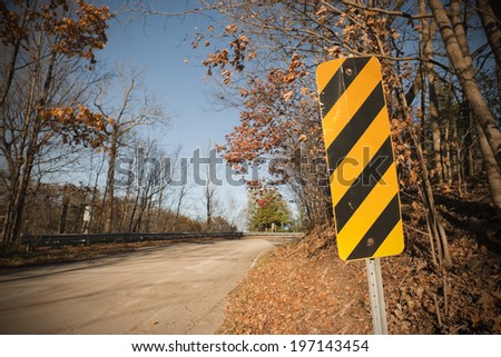 A tree lined country road with a black and yellow sign.