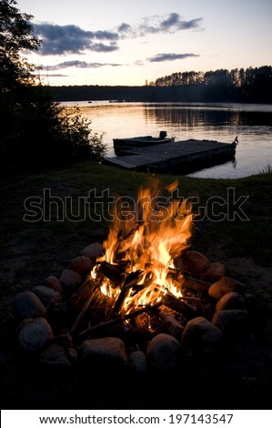 A campfire near a lake, surrounded by trees, with a dock and a boat.