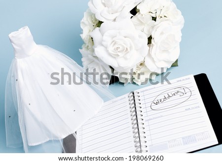 A miniature wedding dress and white bouquet of roses, with a day planner marked Wedding.