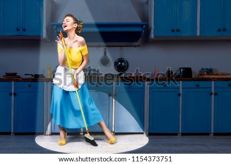 Retro pin up girl woman female housewife wearing yellow top, blue skirt and white apron holding mop singing and cleaning floor in stage light kitchen with blue cabinets and utensils. Housework concept